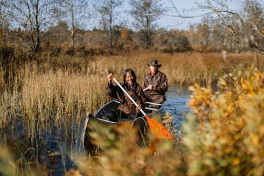 Södamerikansk dam med flätor och en man med en cowboyhatt paddlar kanot på en smal flod omgiven av gräs och natur.
