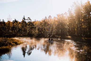 Two people canoeing in a calm river with fog on the water surface while the sun peeking through the trees.