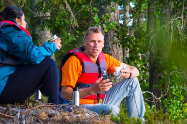 Two people are sitting on a forested hillside, enjoying a meal while wearing life jackets.