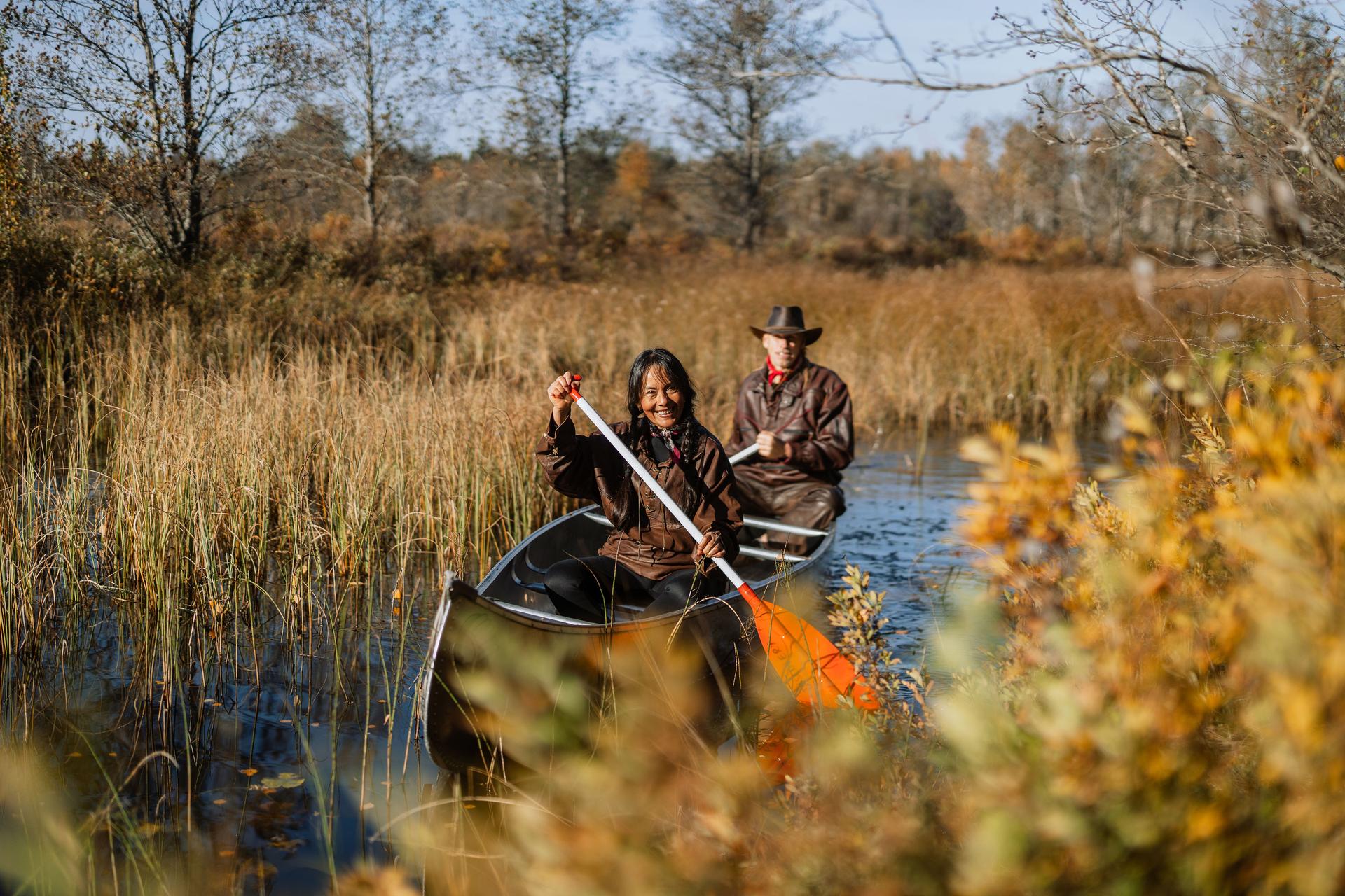 South American lady with braids and a man with a cowboy hat canoeing in a narrow river surrounded by grass and nature.