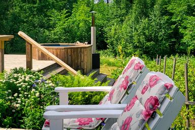 Wood-heated hot tub and seating area in the sunshine surrounded by green trees.