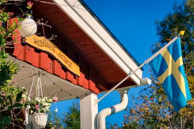 Ein Holzschild mit dem Namen 'Wildnis-Hütte' im Sonnenschein und eine schwedische Flagge, die im Wind weht.