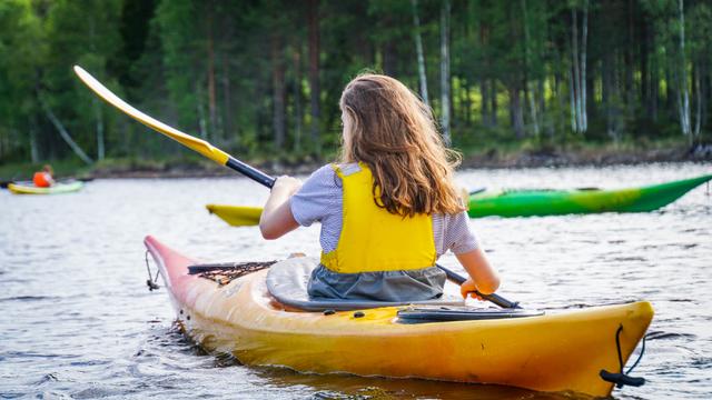 Eine junge Frau ist in einem Kajak auf einem ruhigen See und trägt eine gelbe Schwimmweste. Sie paddelt vorwärts in einem bunten Kajak und im Hintergrund sind Bäume zu sehen.