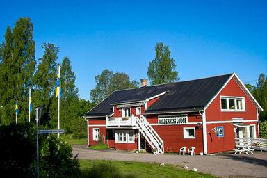 Den traditionella svenska Wilderness Lodge omgiven av träd och grönska. Himmelen är blå utan en enda moln. Tre svenska flaggor i trädgården.