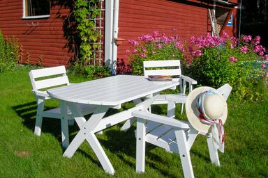 Drei weiße Holzgartenstühle im Garten der wilderness lodge in Schweden. Ein Buch und ein Sonnenhut auf einem der Stühle.