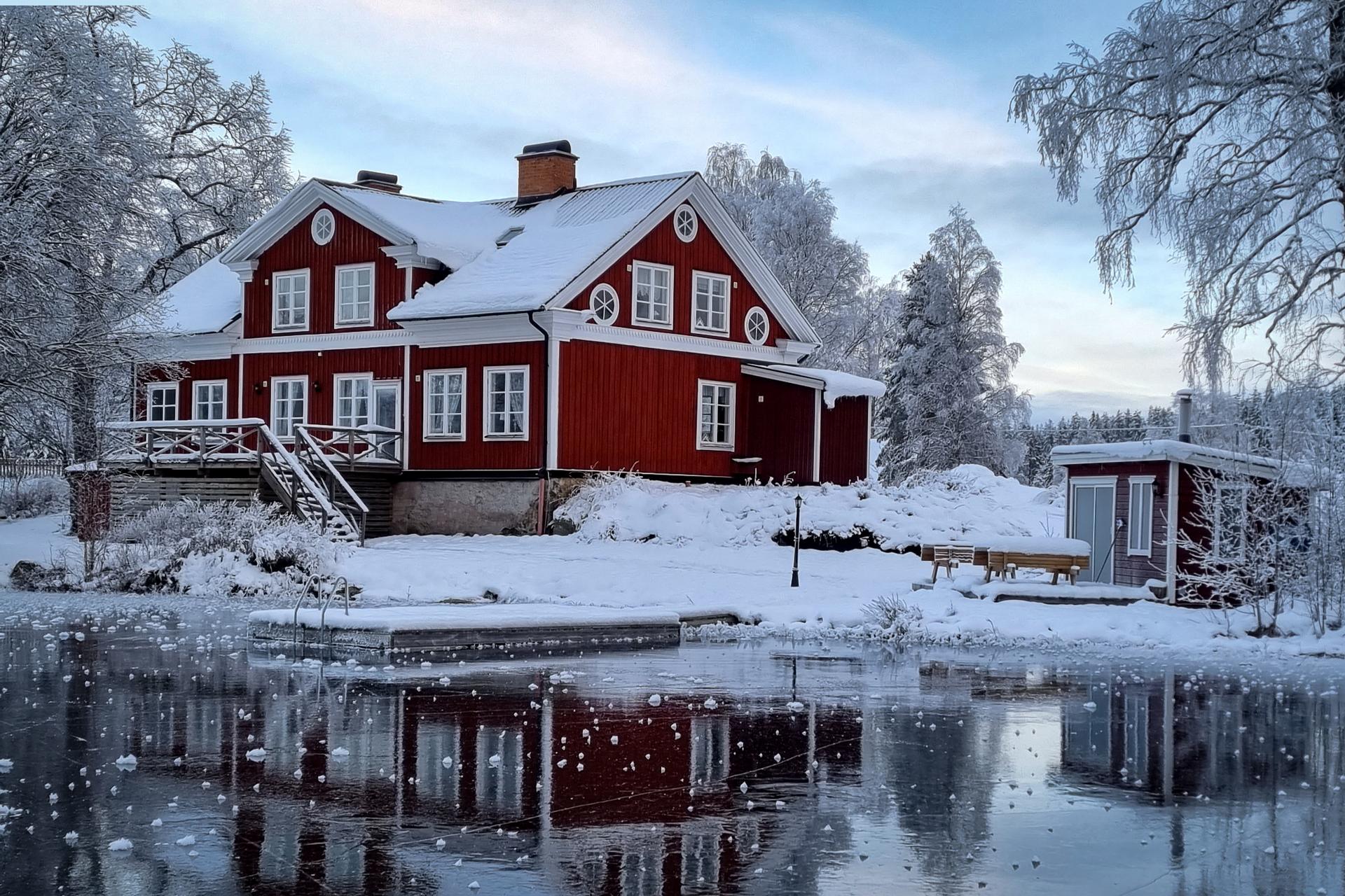 The river lodge is in the middle of a snowy winter landscape with ice on the lake and snow on the trees.