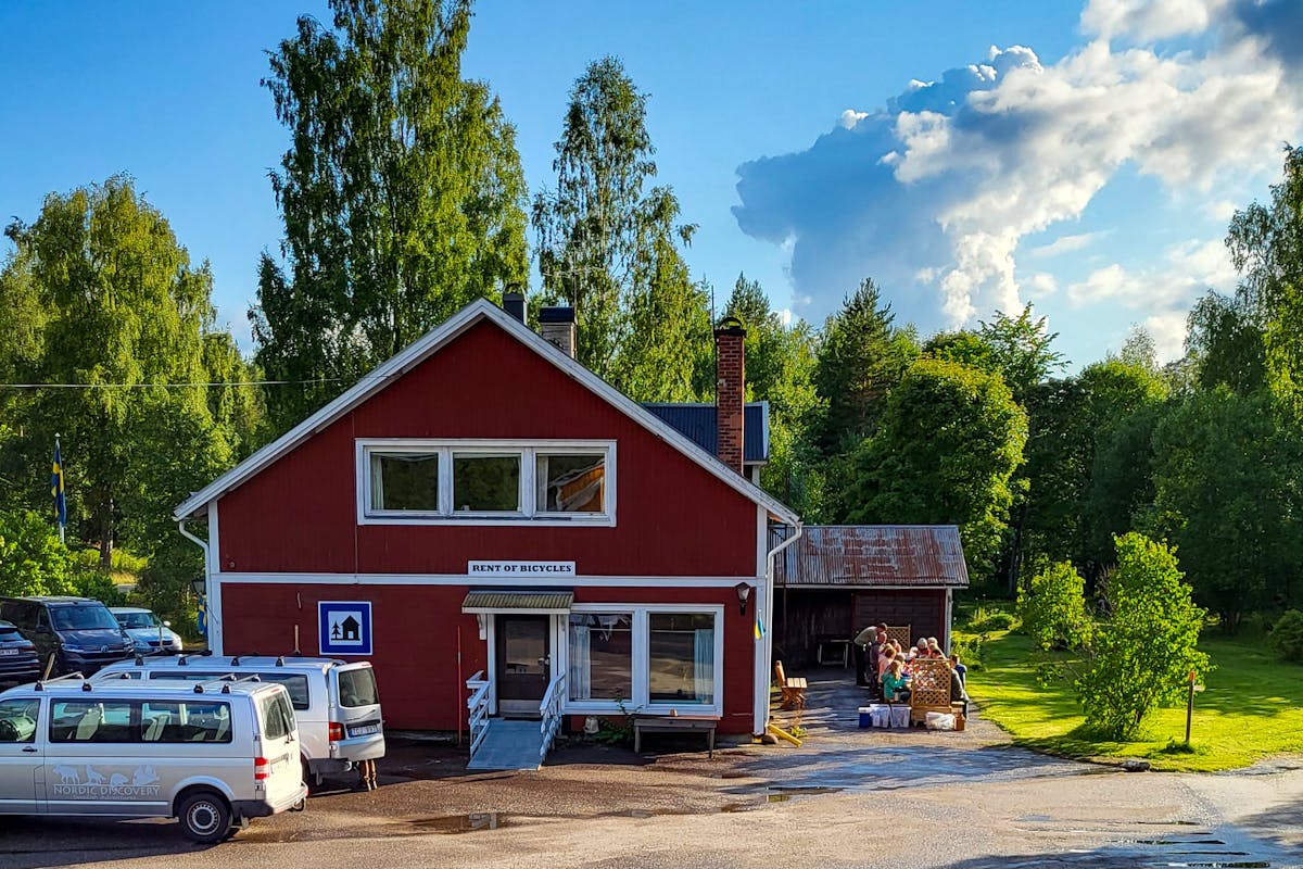 Wilderness Lodge i Sverige med en traditionell röd och vit träfasad. Himmelen är blå med några moln. Trädgården är grön och frodig med människor som äter ute.