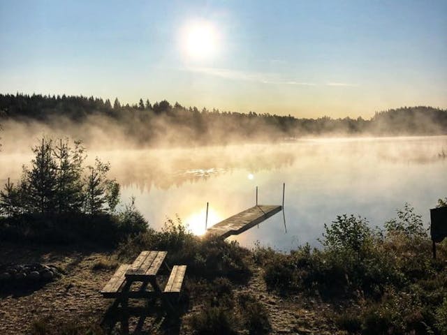 In the early morning at the wilderness camping, smoke rises above the calm water, a jetty extends into the lake, and on the land, there is a fireplace and seating area.